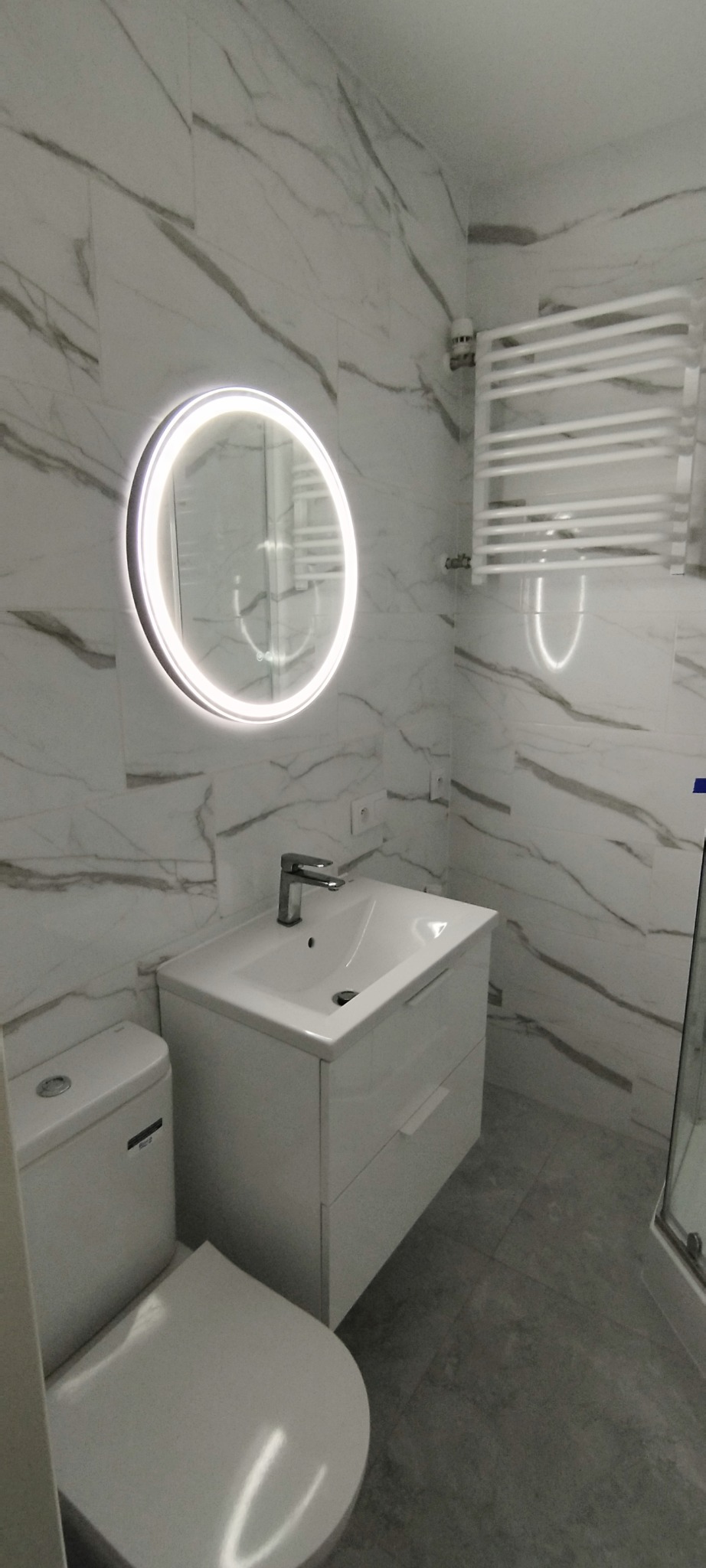 Nowoczesna łazienka z białymi płytkami imitującymi marmur, okrągłym lustrem z podświetleniem LED i grzejnikiem drabinkowym. Umywalka z szafką i toaleta w kolorze białym.