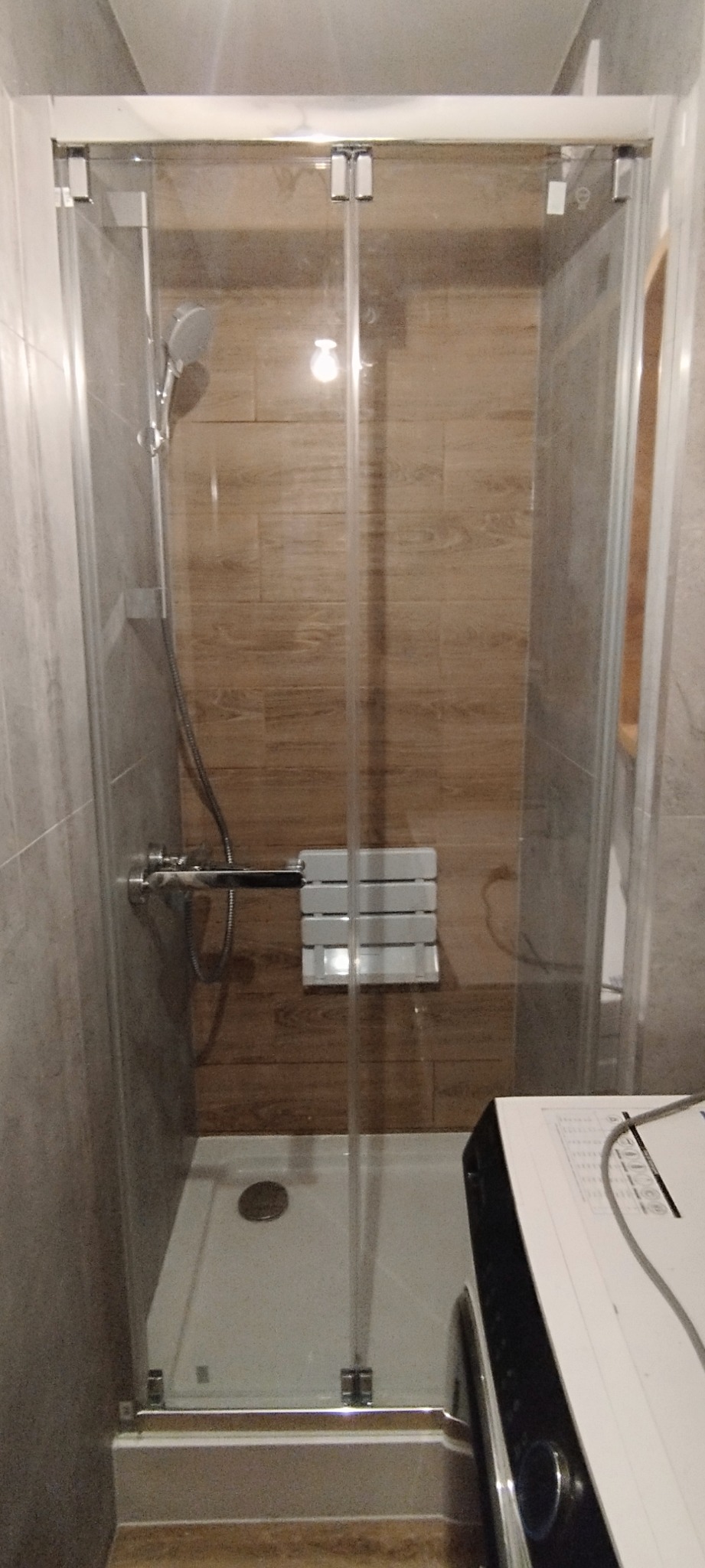 Kabina prysznicowa z transparentnymi drzwiami, chromowanymi zawiasami i siedziskiem. Ściany wyłożone płytkami imitującymi drewno i szarym kamieniem.