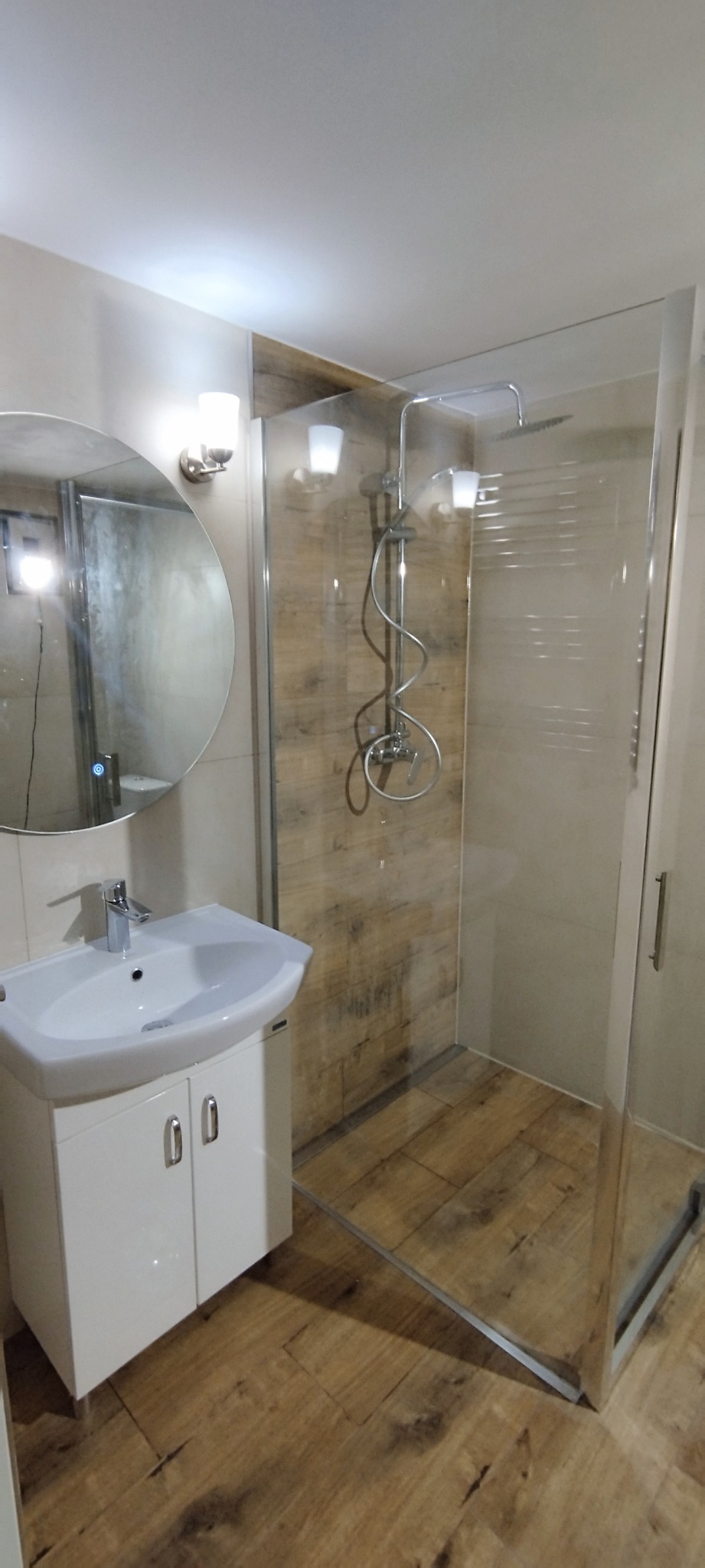 Jasna łazienka po remoncie: kabina prysznicowa ze szklanymi drzwiami, umywalka z szafką, okrągłe lustro z oświetleniem. Płytki imitujące drewno na podłodze i ścianie.