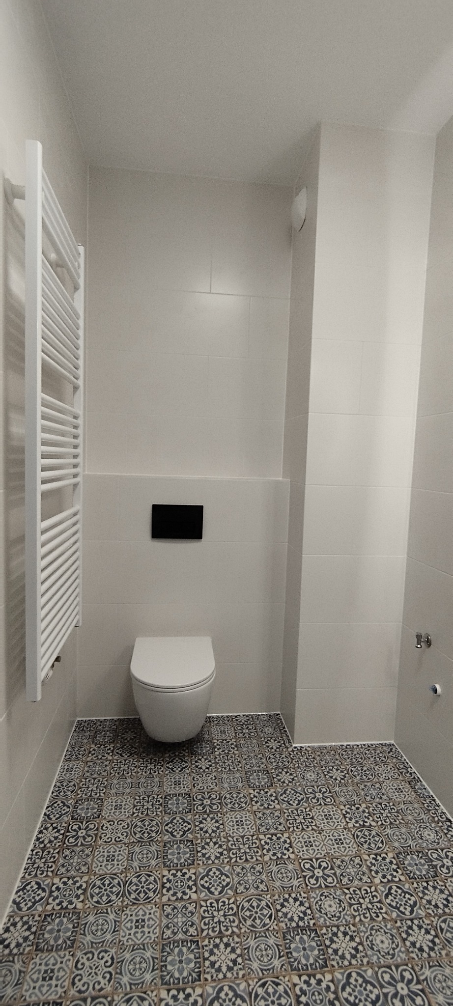 Nowoczesna łazienka z wiszącą toaletą, grzejnikiem drabinkowym i wzorzystą podłogą z płytek. Minimalistyczny design, jasne płytki ścienne i czarny panel sterujący.