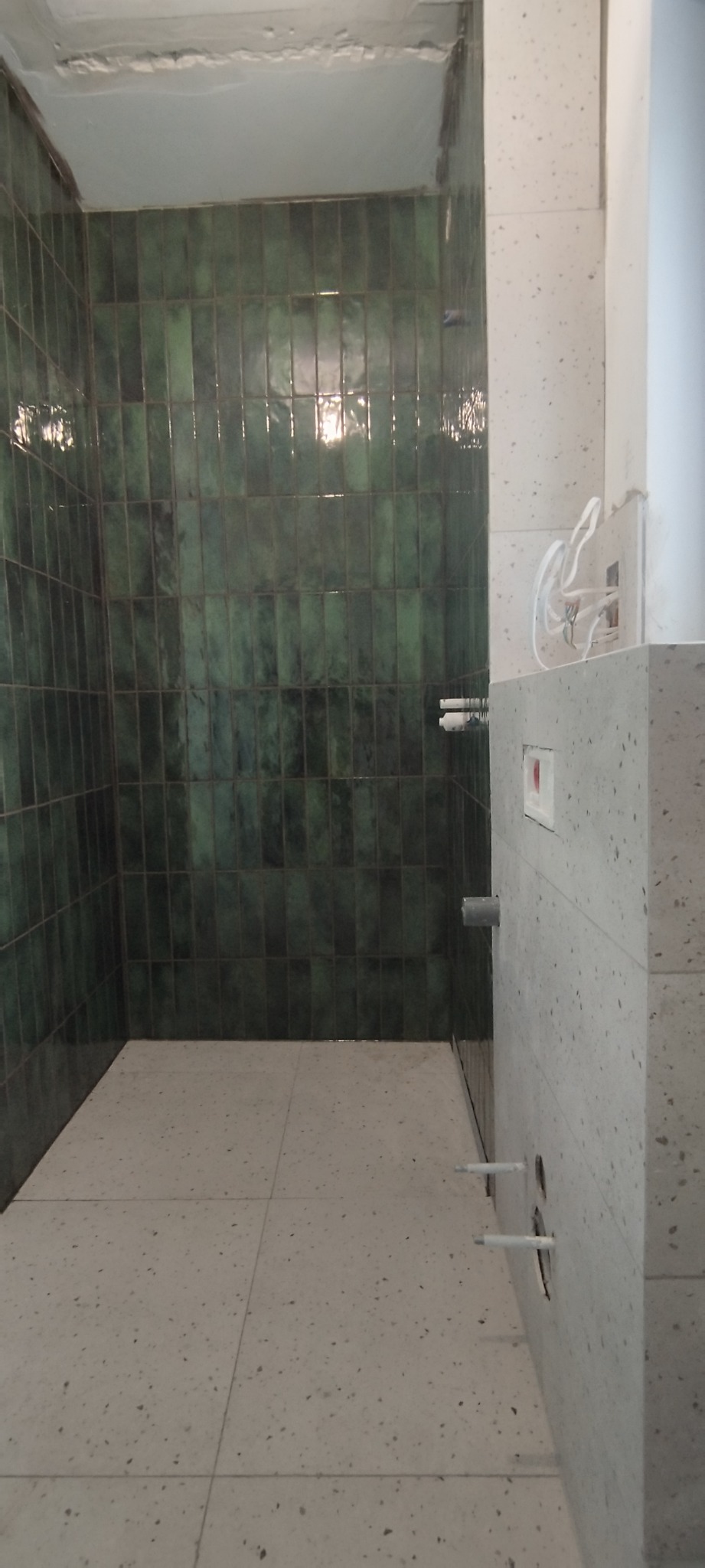 Wykończona łazienka z zielonymi, wąskimi płytkami na ścianach i jasną podłogą w kropki. Widoczne elementy instalacji podtynkowej.