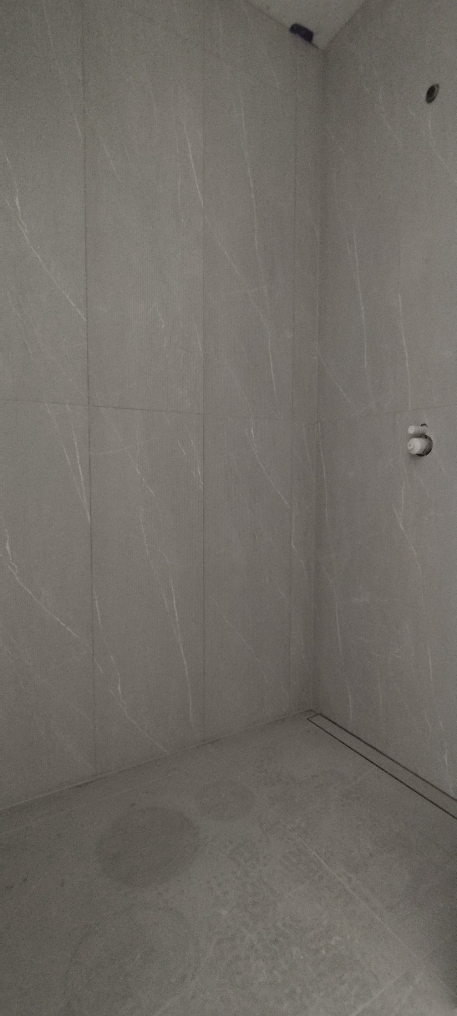 Wykończona kabina prysznicowa z szarymi płytkami imitującymi marmur z delikatnym wzorem żyłkowania. Widoczny odpływ liniowy i otwór na baterię.