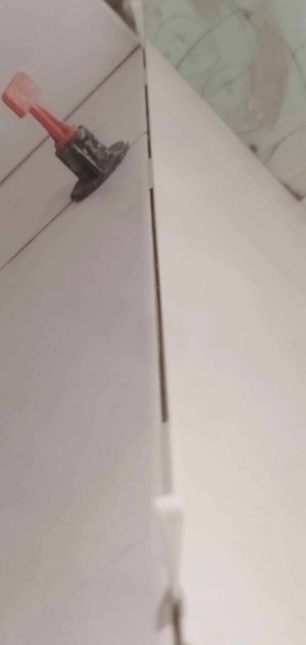 Białe płytki ścienne z systemem poziomowania, widok z bliska na narożnik. Cienka fuga. Czerwony klin i czarna baza systemu poziomowania płytek.