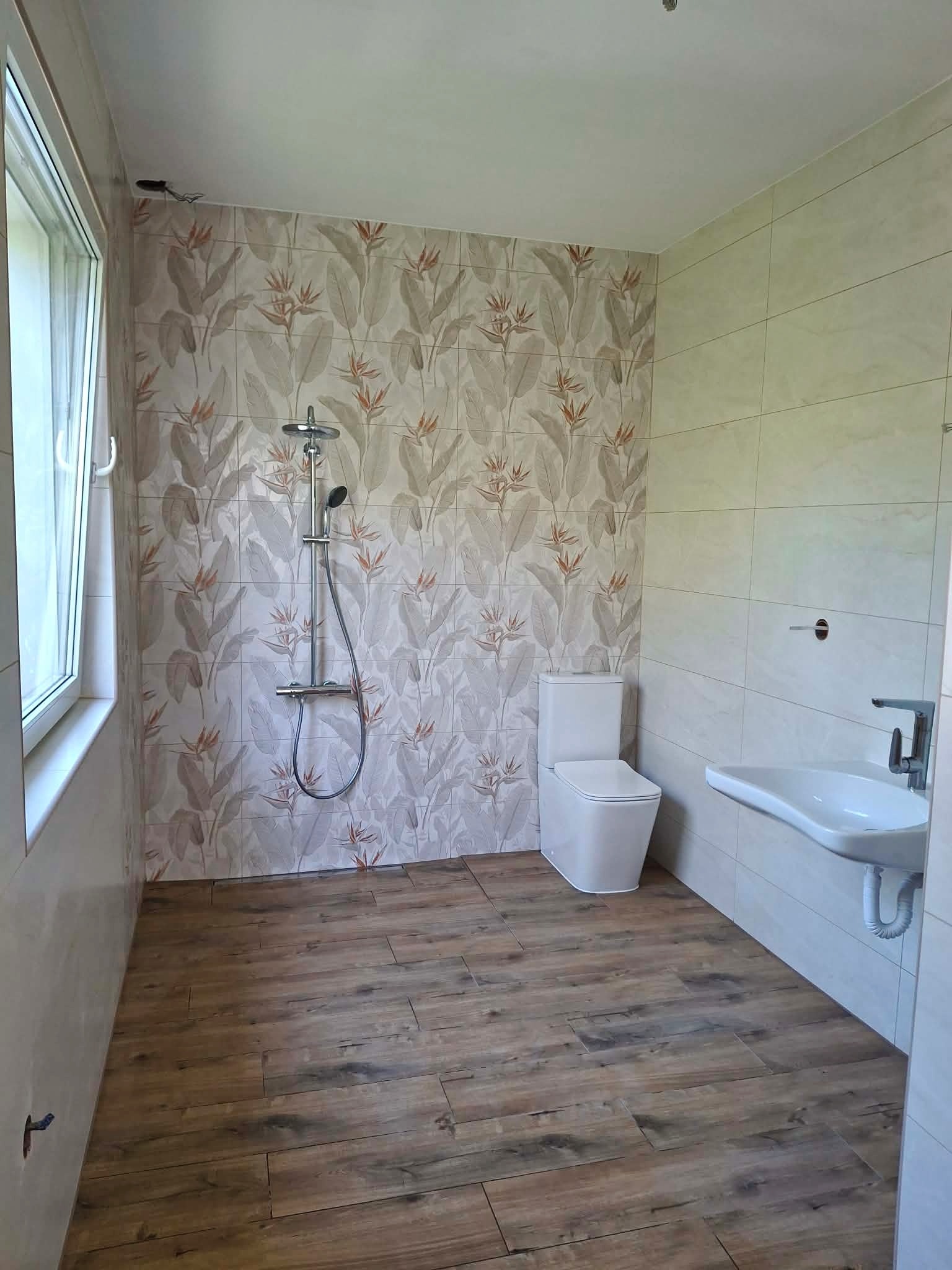 Jasna łazienka z drewnopodobną podłogą, wzorzystymi płytkami ściennymi z motywem roślinnym, białą armaturą i kabiną prysznicową. Nowoczesny design.