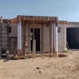 Magdalena Szczepańska BUDIMAG - Murowany budynek w trakcie budowy, widoczne bloczki, drewniane szalunki i podpory, worki z zaprawą obok drabiny.