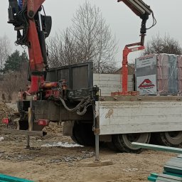 Magdalena Szczepańska BUDIMAG - Ciężarówka z czerwoną dźwigiem hydraulicznym Fassi, załadowana paletami dachówek Meyer-Holsen, na placu budowy z zielonymi belkami w pierwszym planie.