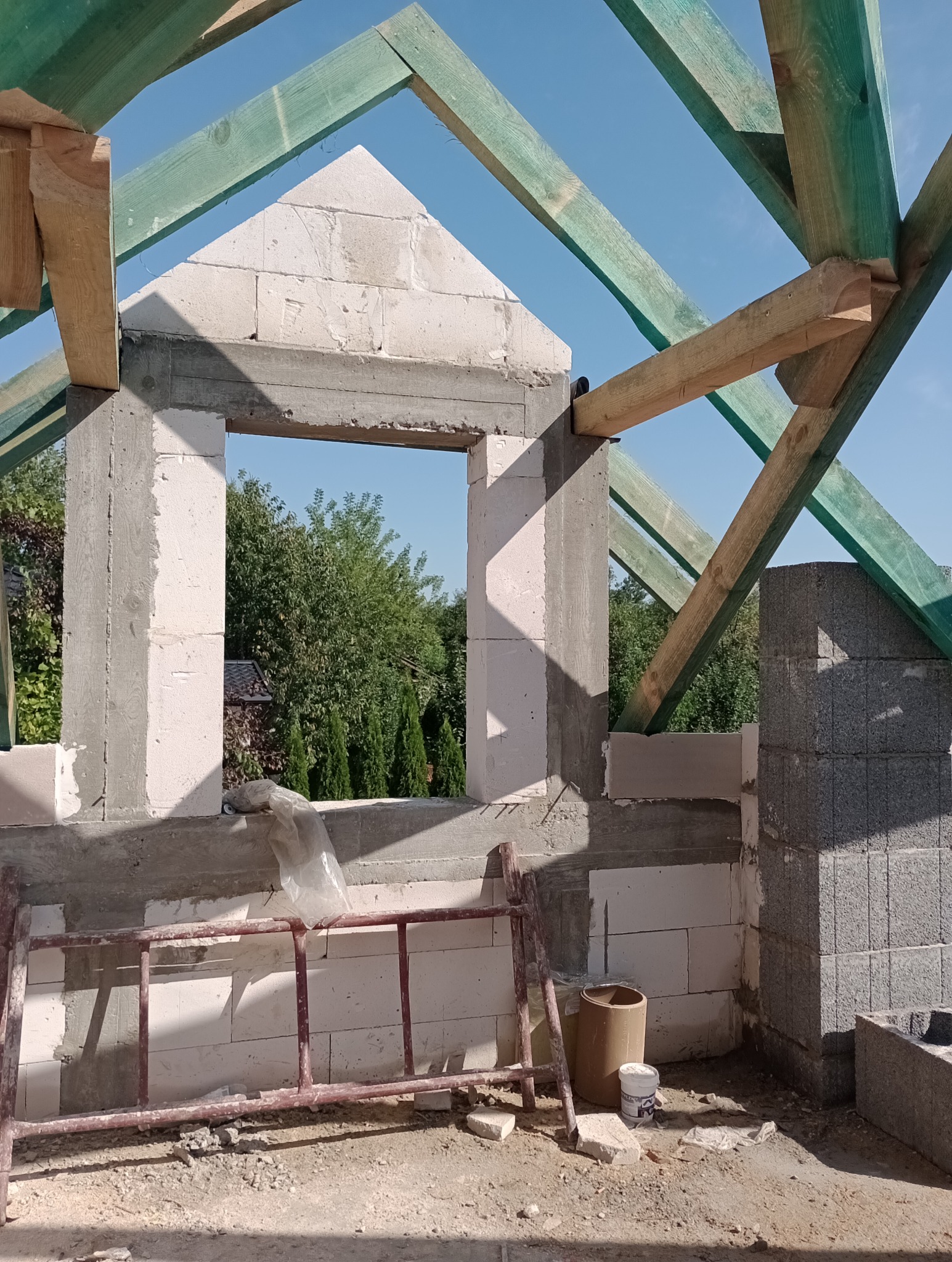 Wznoszona ściana budynku z pustaków, widoczne drewniane elementy konstrukcji dachu, drabina i pojemniki na zaprawę murarską na placu budowy.