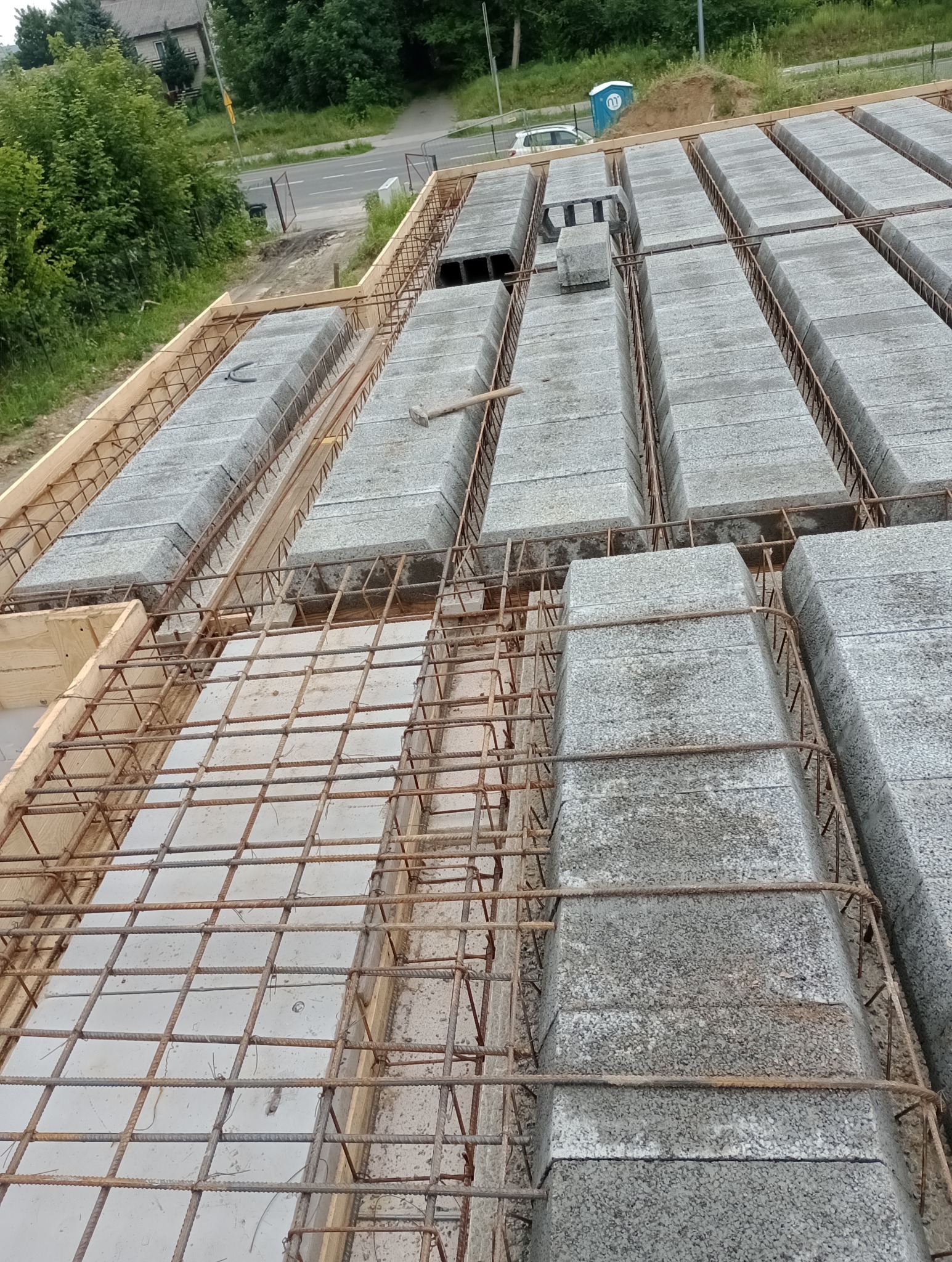 Szary strop Teriva w trakcie montażu z widoczną siatką zbrojeniową i pustakami betonowymi; w tle widoczny teren budowy z przenośną toaletą.