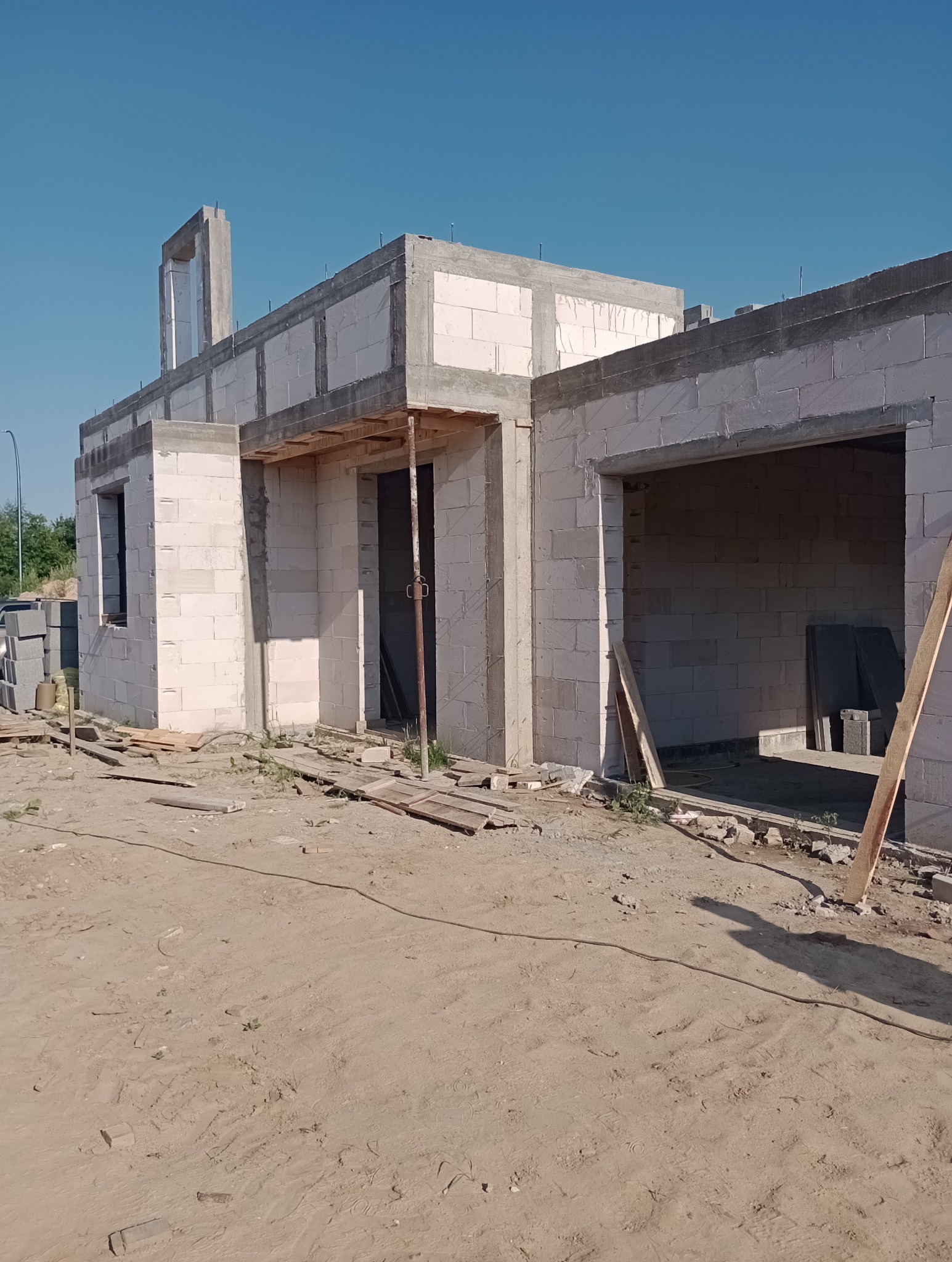 Surowy stan budowy domu jednorodzinnego z białych bloczków, widoczne przygotowane otwory okienne i drzwiowe, teren wokół niezagospodarowany, słoneczny dzień.