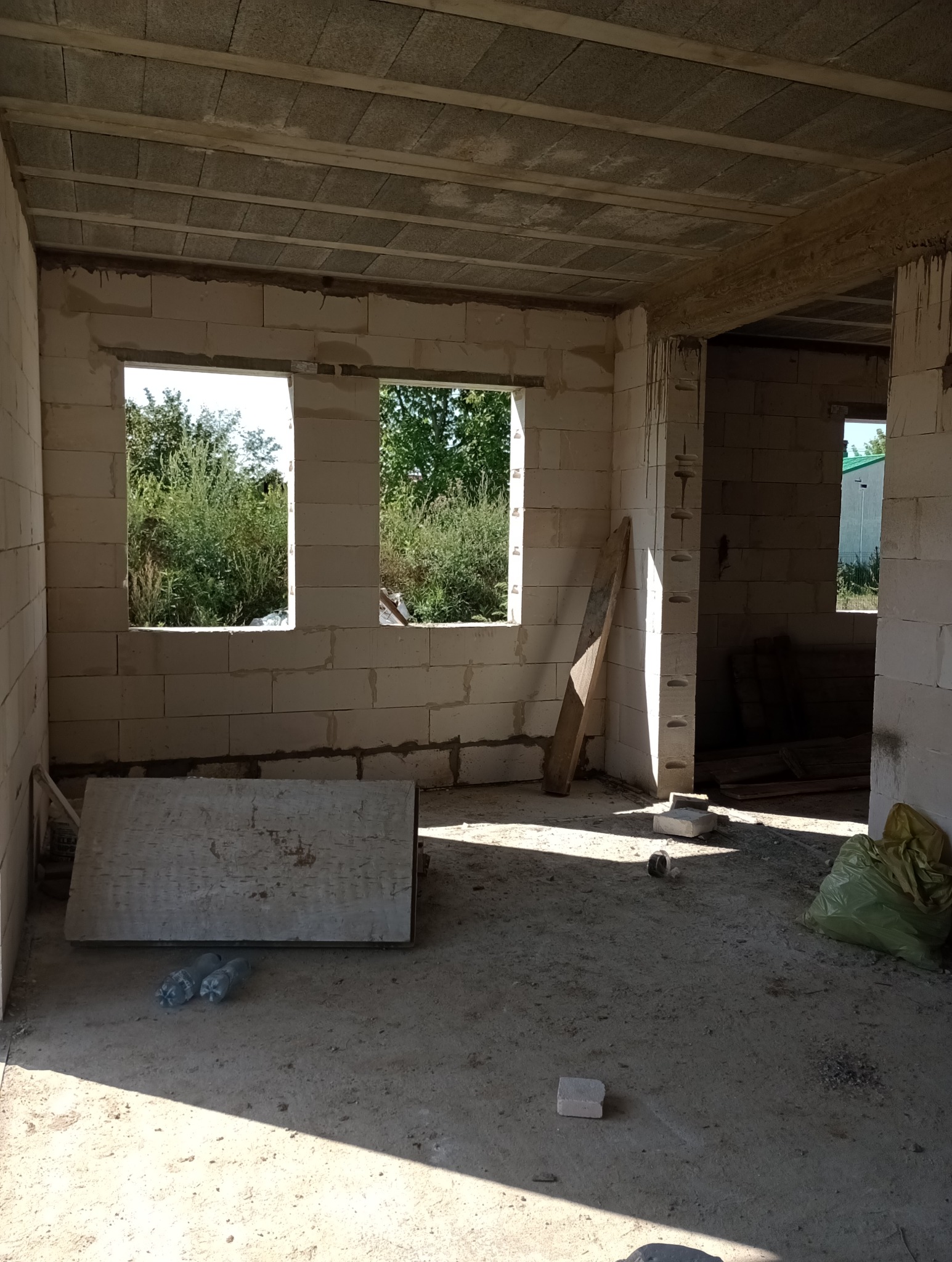 Wnętrze surowego domu w trakcie budowy, widoczne ściany z białych bloczków, otwory okienne z widokiem na zieleń, betonowy strop, deska oparta o ścianę, butelki i worek na podłodze.