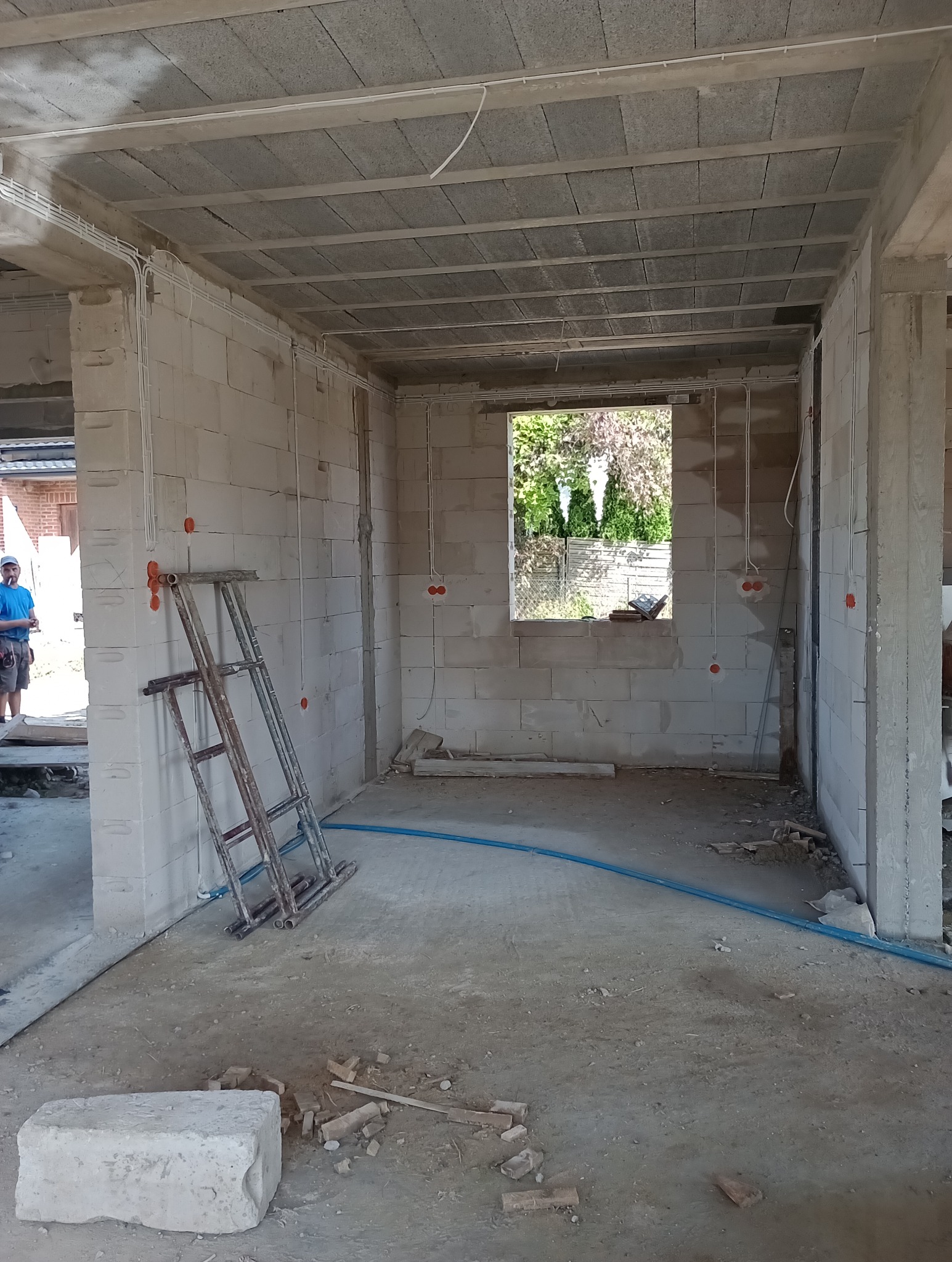 Surowy stan wnętrza domu w trakcie budowy: ściany z białych bloczków, betonowy strop, instalacja elektryczna w peszlach, drabina oparta o ścianę, widok przez okno na zieleń.