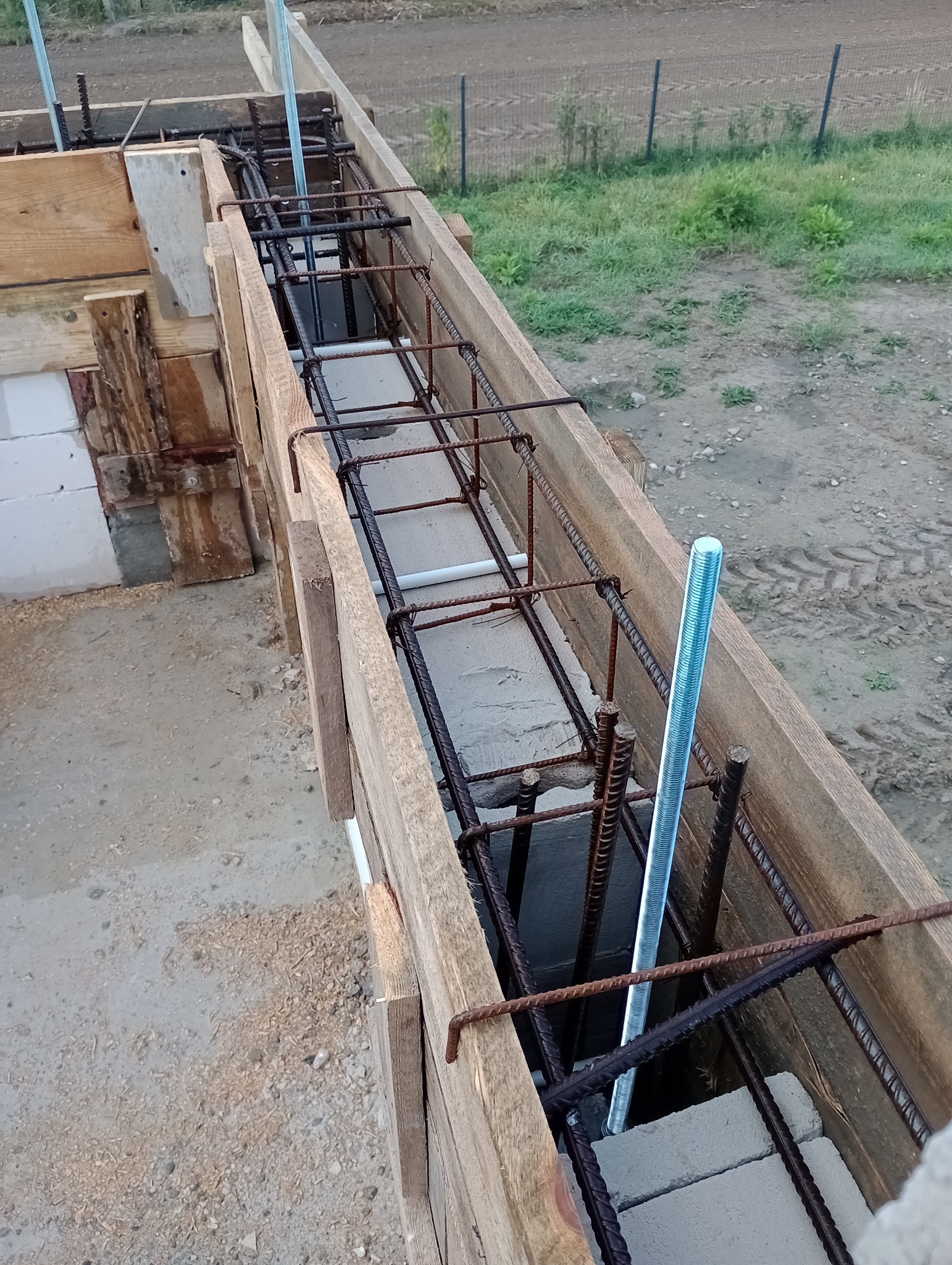 Zbrojenie fundamentu w drewnianym szalunku, widoczne wystające pręty gwintowane, bloczki betonowe i fragment ściany z białych pustaków.