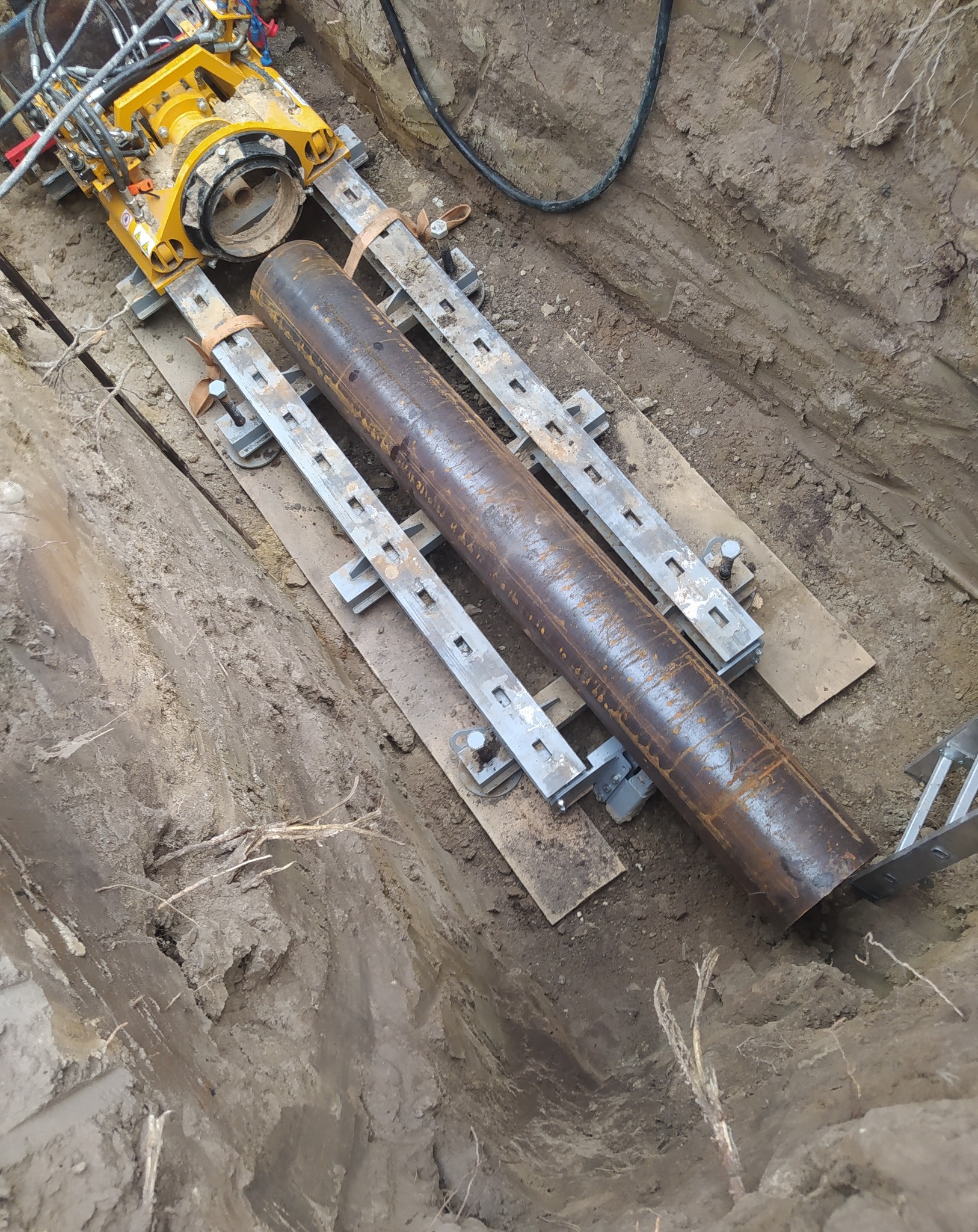 Żółta maszyna do przewiertów sterowanych, zamontowana na szynach w wykopie, przygotowuje się do wciągnięcia rury stalowej.