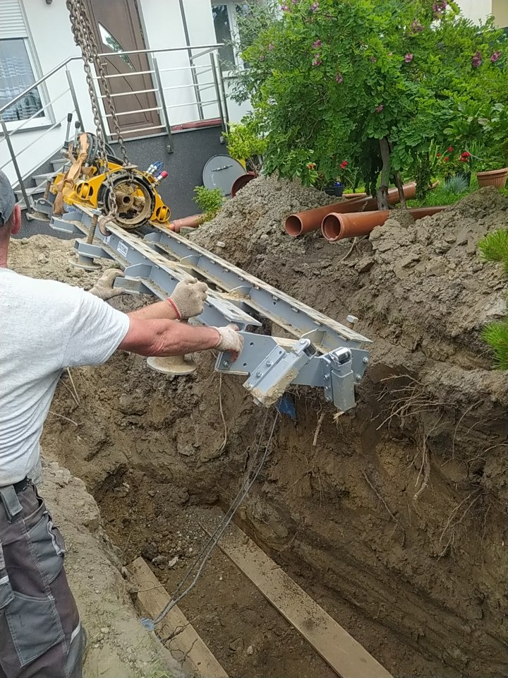 Żółta maszyna do przewiertów sterowanych opuszczana do wykopu za pomocą łańcucha, widoczne rury kanalizacyjne i fragment budynku mieszkalnego z ogrodem w tle.