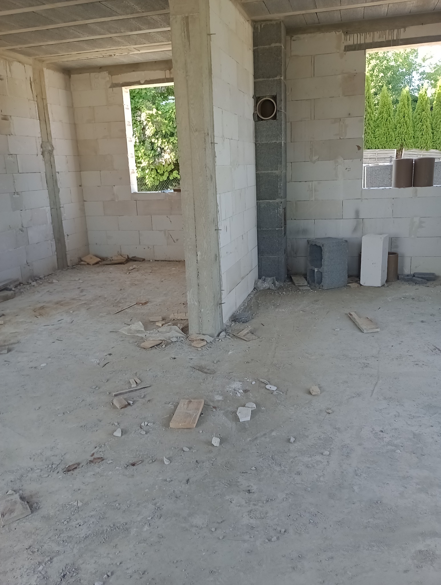 Surowy stan wnętrza budynku w trakcie budowy: ściany z bloczków, betonowy strop, otwory okienne z widokiem na zieleń, widoczny przewód wentylacyjny w ścianie.