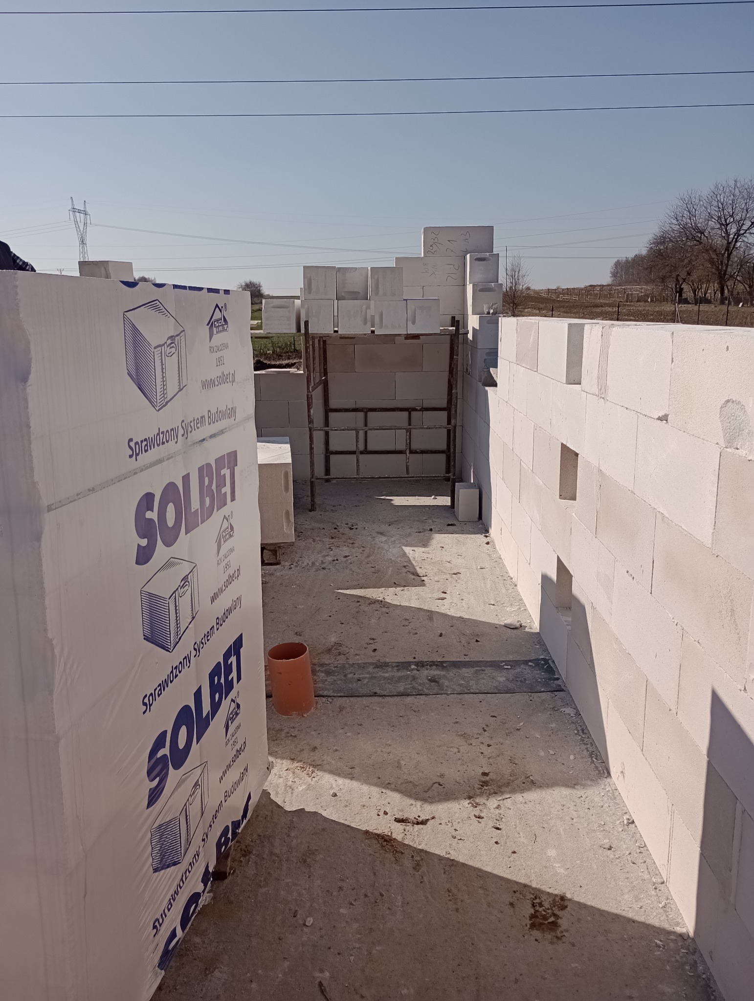 Wznoszenie ścian z białych bloczków Solbet na budowie, widoczne palety z materiałem i fragment konstrukcji rusztowania w tle, w słoneczny dzień.