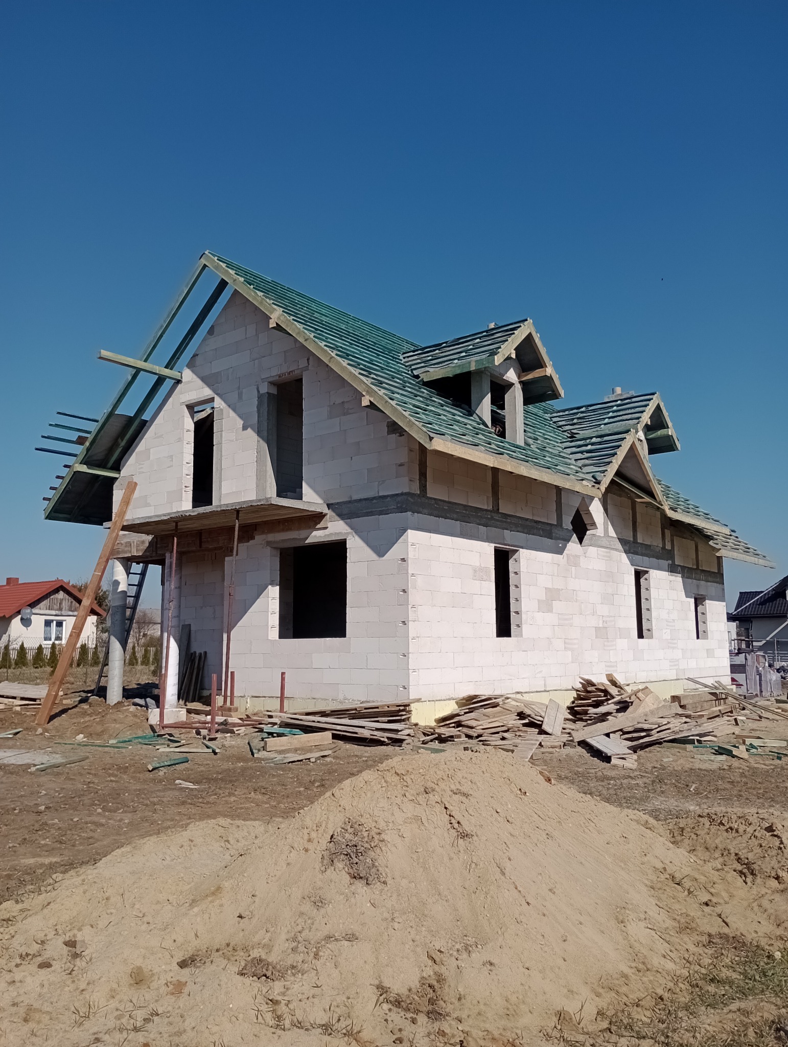Niedokończony dom jednorodzinny z białego bloczka, widoczne zarysowane okna i dach z zieloną membraną, na pierwszym planie hałda piasku i porozrzucane deski.
