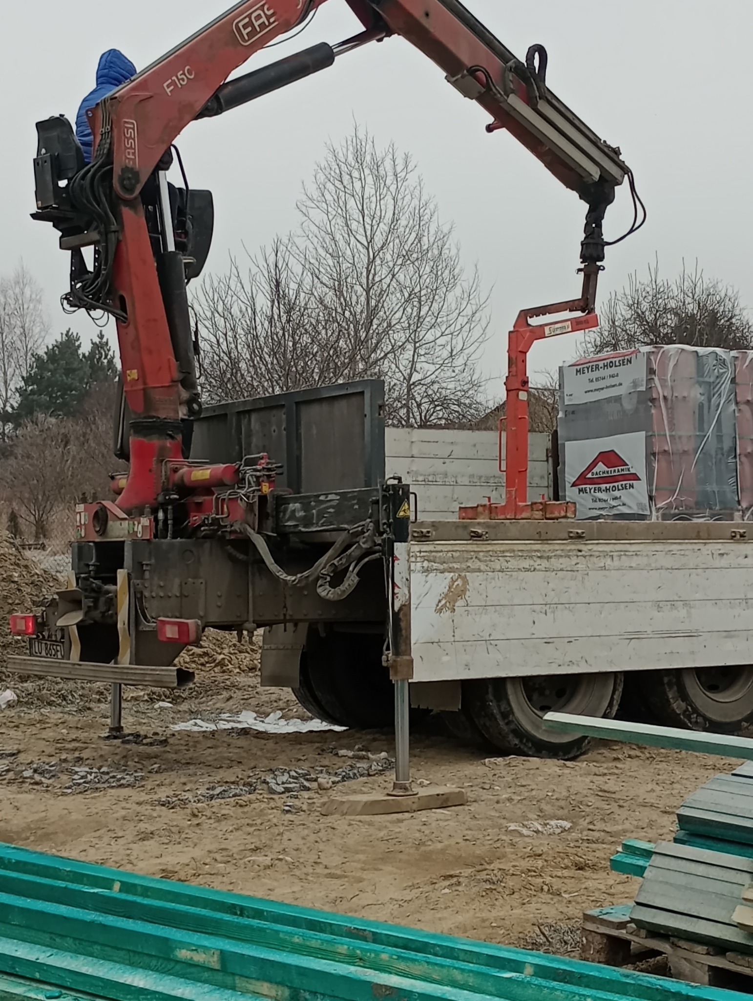 Ciężarówka z czerwoną dźwigiem hydraulicznym Fassi, załadowana paletami dachówek Meyer-Holsen, na placu budowy z zielonymi belkami w pierwszym planie.