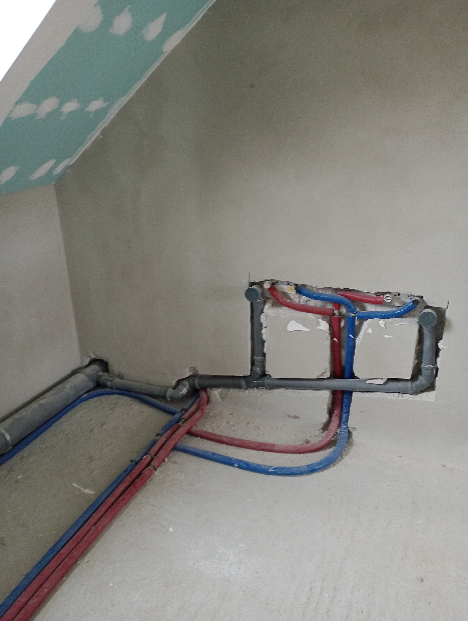 Instalacja wodna i kanalizacyjna w trakcie montażu w surowym wnętrzu, widoczne rury w kolorze niebieskim i czerwonym oraz szare rury kanalizacyjne, ściany w stanie deweloperskim.