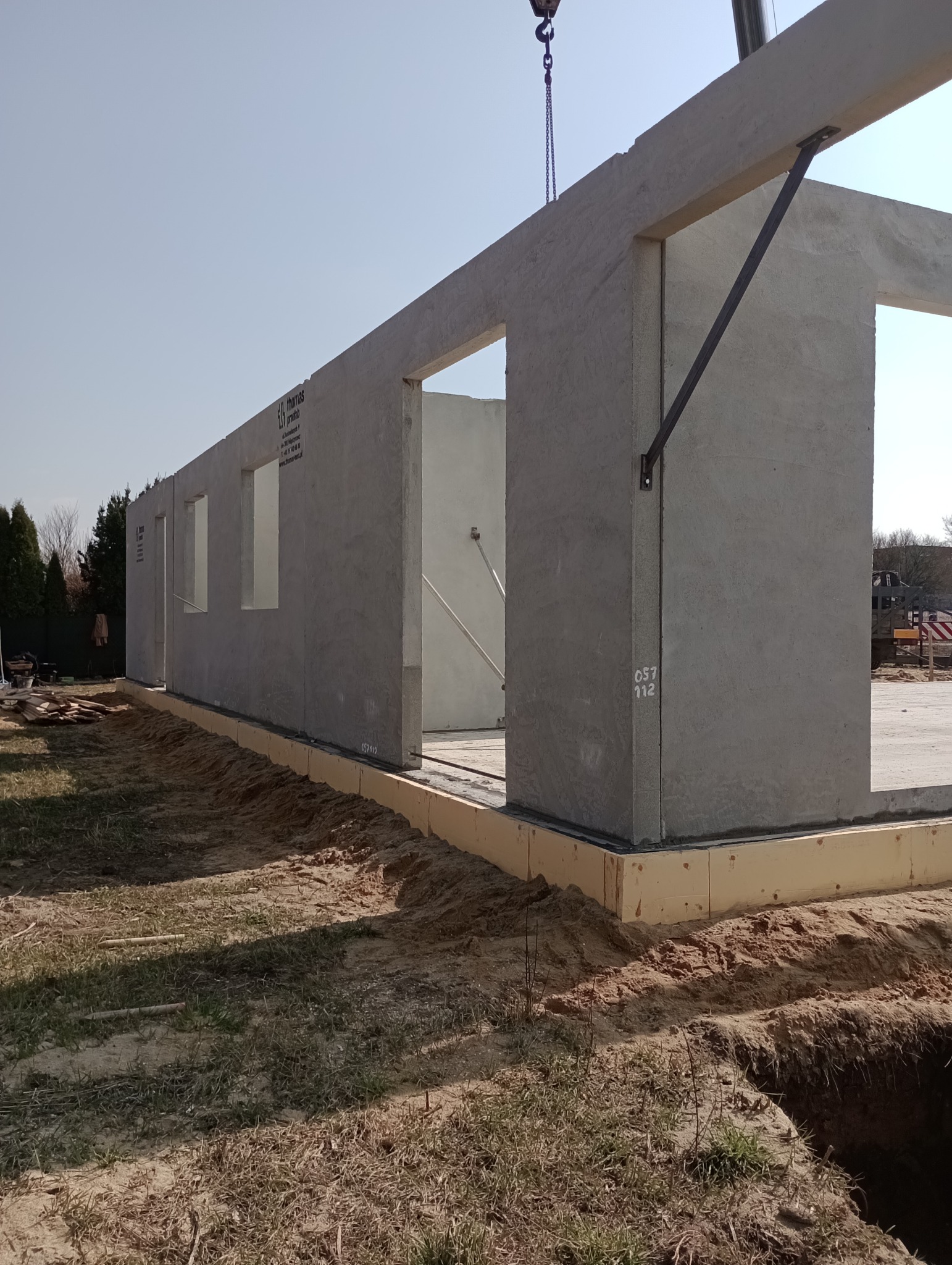 Montaż prefabrykowanych ścian betonowych na fundamencie z izolacją, widoczny dźwig i oznaczenia na ścianie.