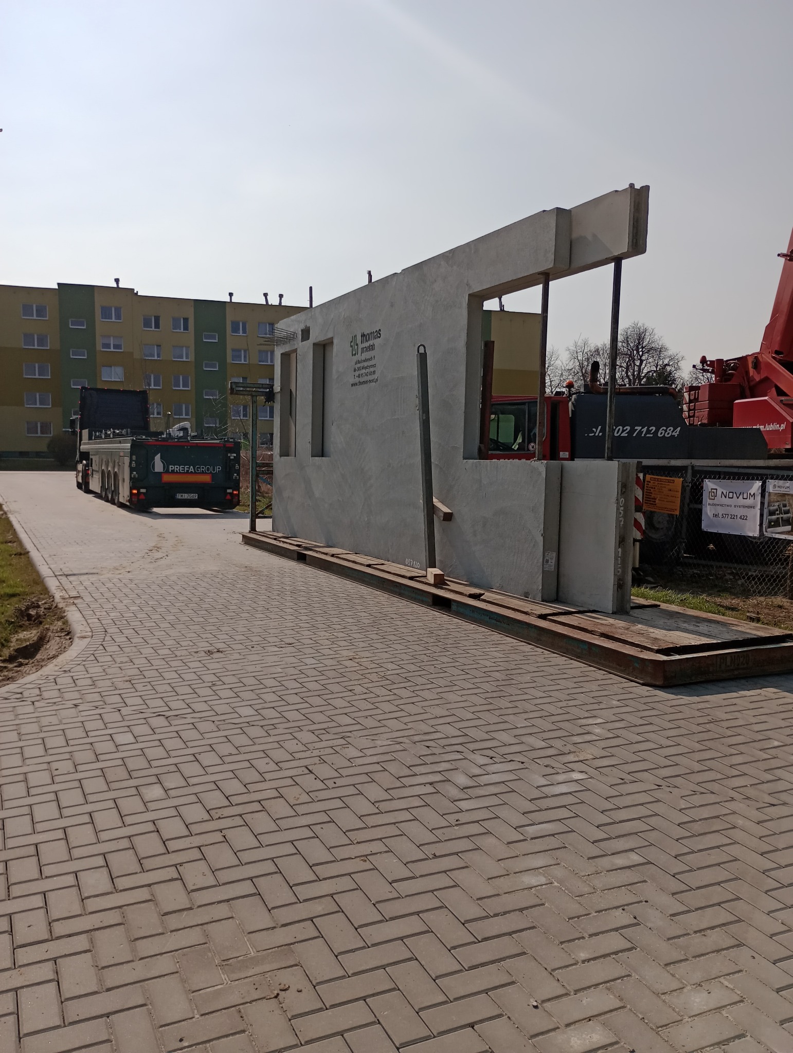 Transport prefabrykowanego betonowego elementu budynku z otworami okiennymi na platformie transportowej; w tle budynek mieszkalny i dźwig budowlany.