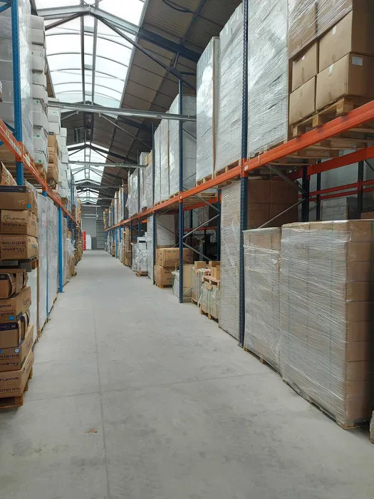 Długi korytarz magazynu z regałami wypełnionymi zapakowanymi towarami, widok w perspektywie z niskiego kąta, oświetlenie górne.