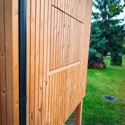 LEKO DESIGN Filip Czuluk - Fragment ściany zewnętrznej budynku obłożonej pionowymi deskami z jasnego drewna, z widoczną czarną rurą spustową i elementem konstrukcyjnym przypominającym okiennicę, na tle zielonego trawnika...