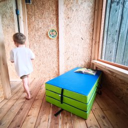 LEKO DESIGN Filip Czuluk - Wnętrze małego domku z drewnianą podłogą i ścianami z płyty OSB, z materacem gimnastycznym w rogu i chłopcem wchodzącym do środka.