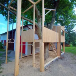Drewniana konstrukcja domku na drzewie z elementami placu zabaw, w tym drabinka, kółka gimnastyczne i zjeżdżalnia linowa, otoczona piaskiem. Konstrukcja wkomponowana w pień drzewa, z widoczną...