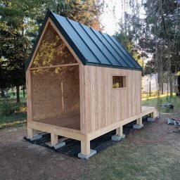 LEKO DESIGN Filip Czuluk - Surowa konstrukcja drewniana małego domku z dachem z blachy na rąbek, posadowiona na betonowych bloczkach, widoczne narzędzia i przewody elektryczne sugerują etap budowy.