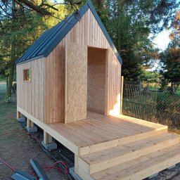 LEKO DESIGN Filip Czuluk - Surowy, drewniany domek z czarnym dachem, posadowiony na betonowych bloczkach, z widocznymi schodami wejściowymi i częściowo zamontowanymi drzwiami z płyty OSB, otoczony zielenią i narzędziami...