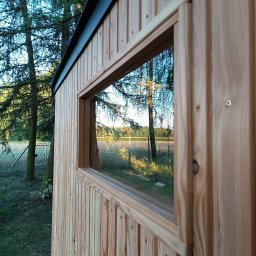 LEKO DESIGN Filip Czuluk - Fragment ściany zewnętrznej domu z drewnianych paneli z widocznym oknem, w którym odbija się krajobraz z drzewami i polem.