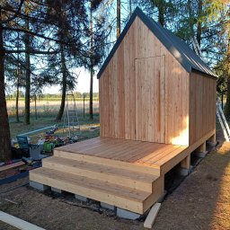 LEKO DESIGN Filip Czuluk - Niewielki, drewniany budynek z dwuspadowym dachem, posadowiony na betonowych bloczkach, z dobudowanym tarasem i schodami wejściowymi, otoczony drzewami i narzędziami budowlanymi.