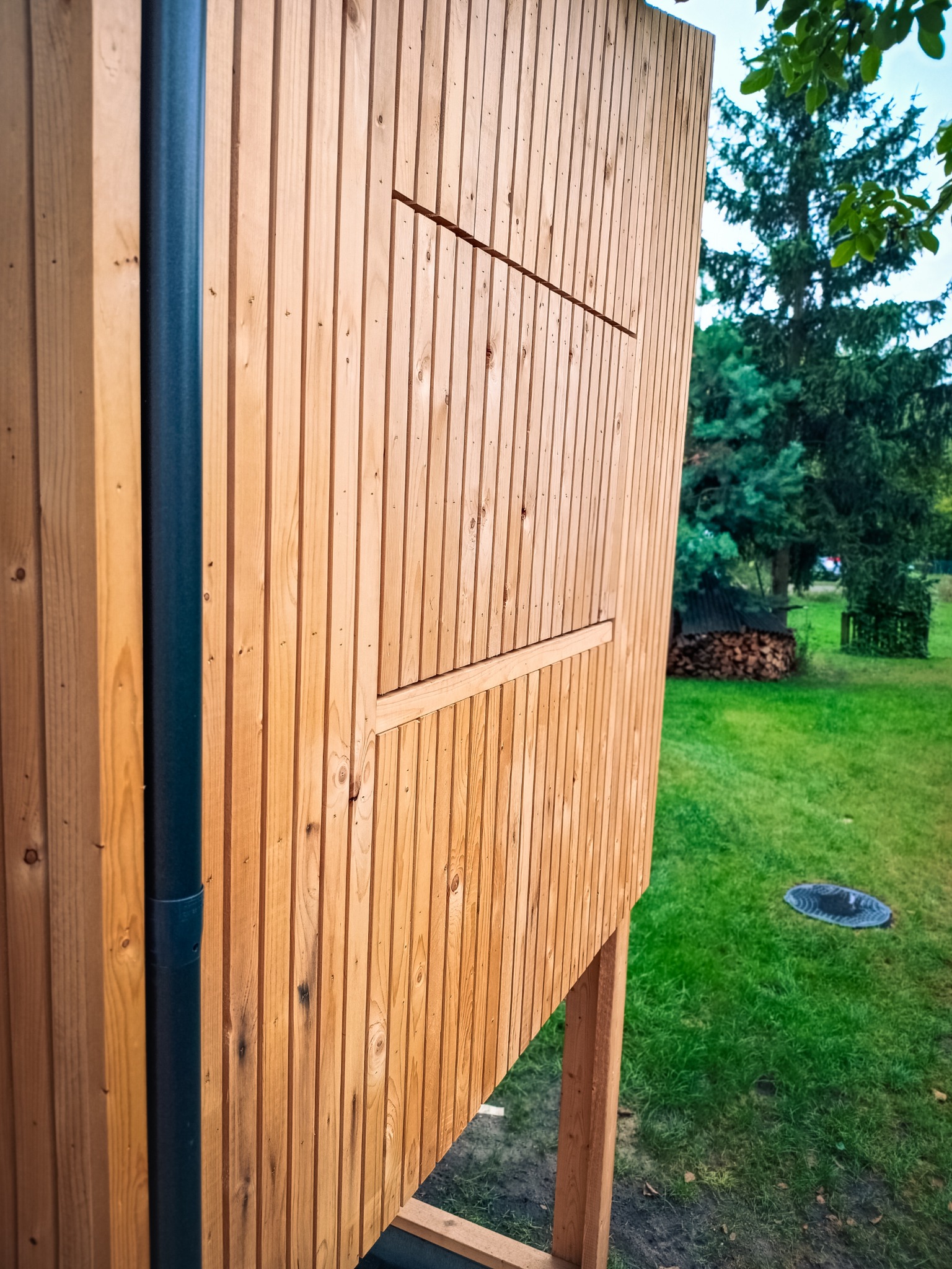 Fragment ściany zewnętrznej budynku obłożonej pionowymi deskami z jasnego drewna, z widoczną czarną rurą spustową i elementem konstrukcyjnym przypominającym okiennicę, na tle zielonego trawnika...