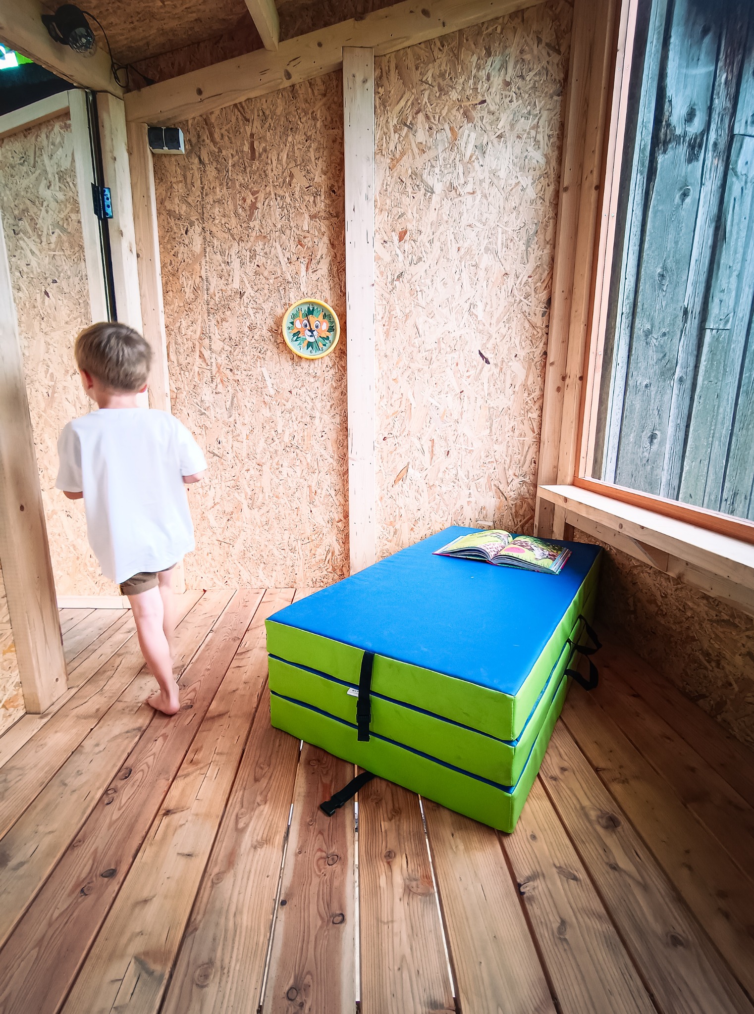 Wnętrze małego domku z drewnianą podłogą i ścianami z płyty OSB, z materacem gimnastycznym w rogu i chłopcem wchodzącym do środka.