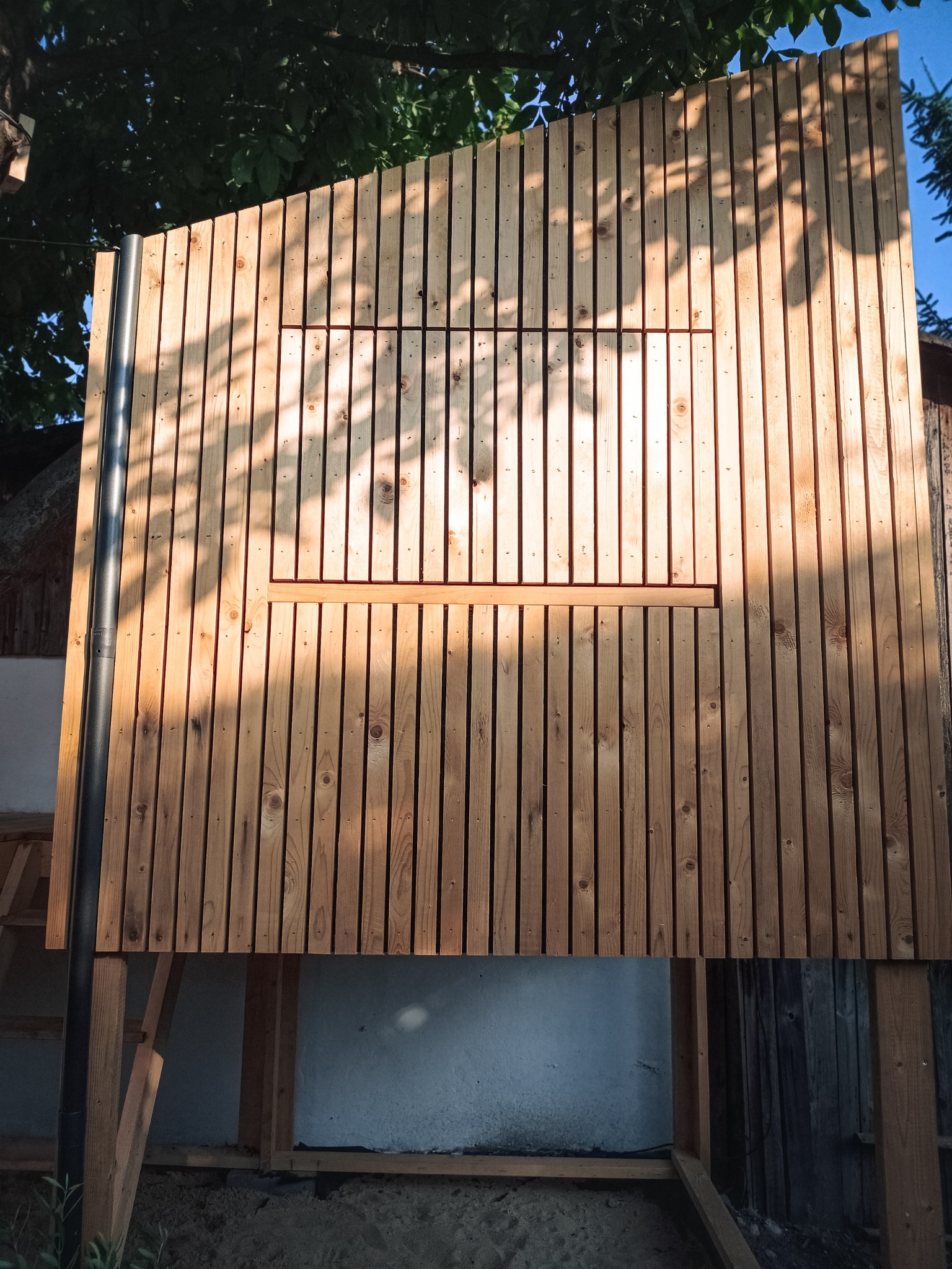 Nowoczesna konstrukcja z pionowych desek z przesłoniętym oknem, oświetlona promieniami słońca, z widoczną czarną rurą i drewnianymi schodkami z boku.