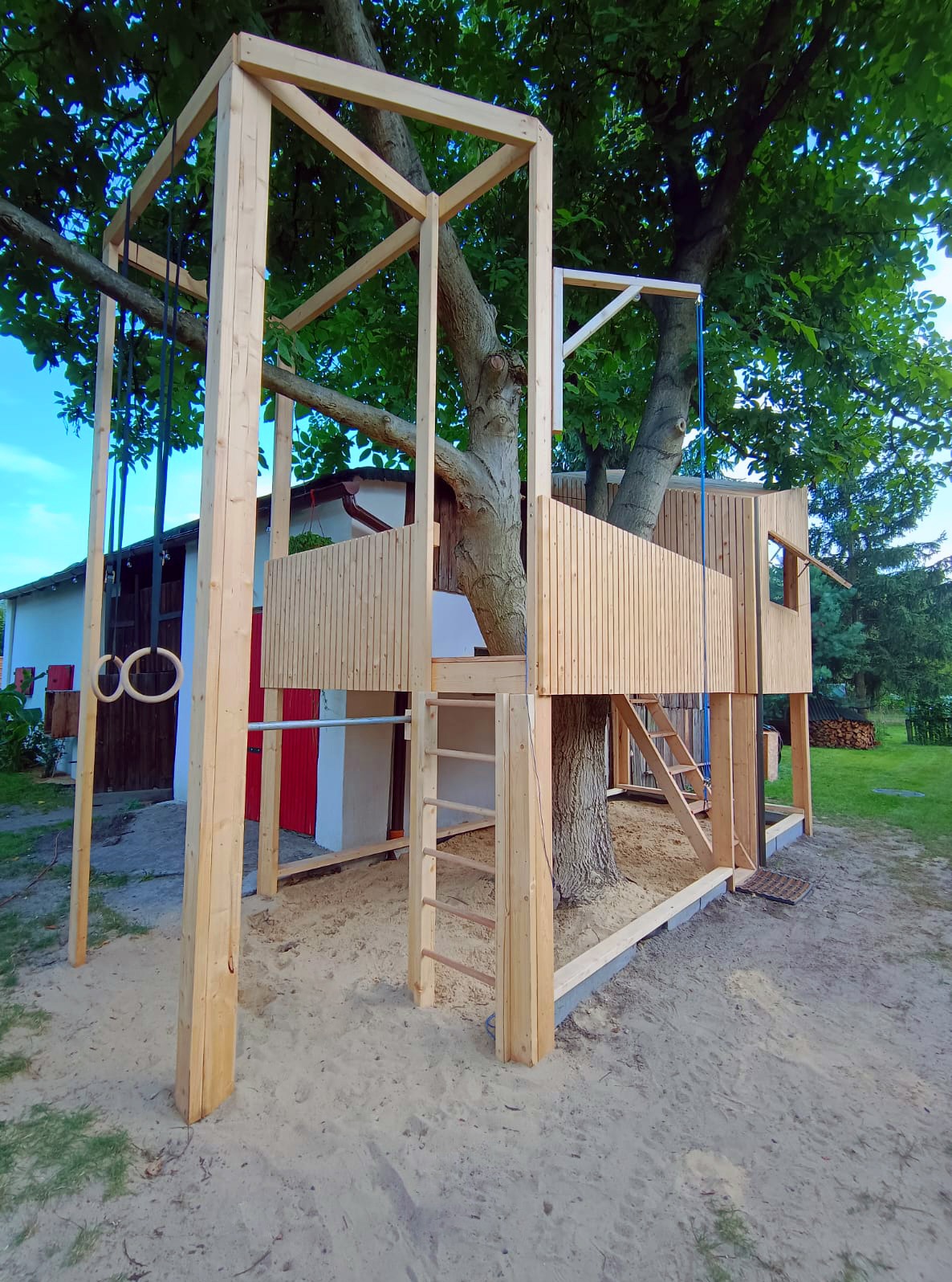Drewniana konstrukcja domku na drzewie z elementami placu zabaw, w tym drabinka, kółka gimnastyczne i zjeżdżalnia linowa, otoczona piaskiem. Konstrukcja wkomponowana w pień drzewa, z widoczną...