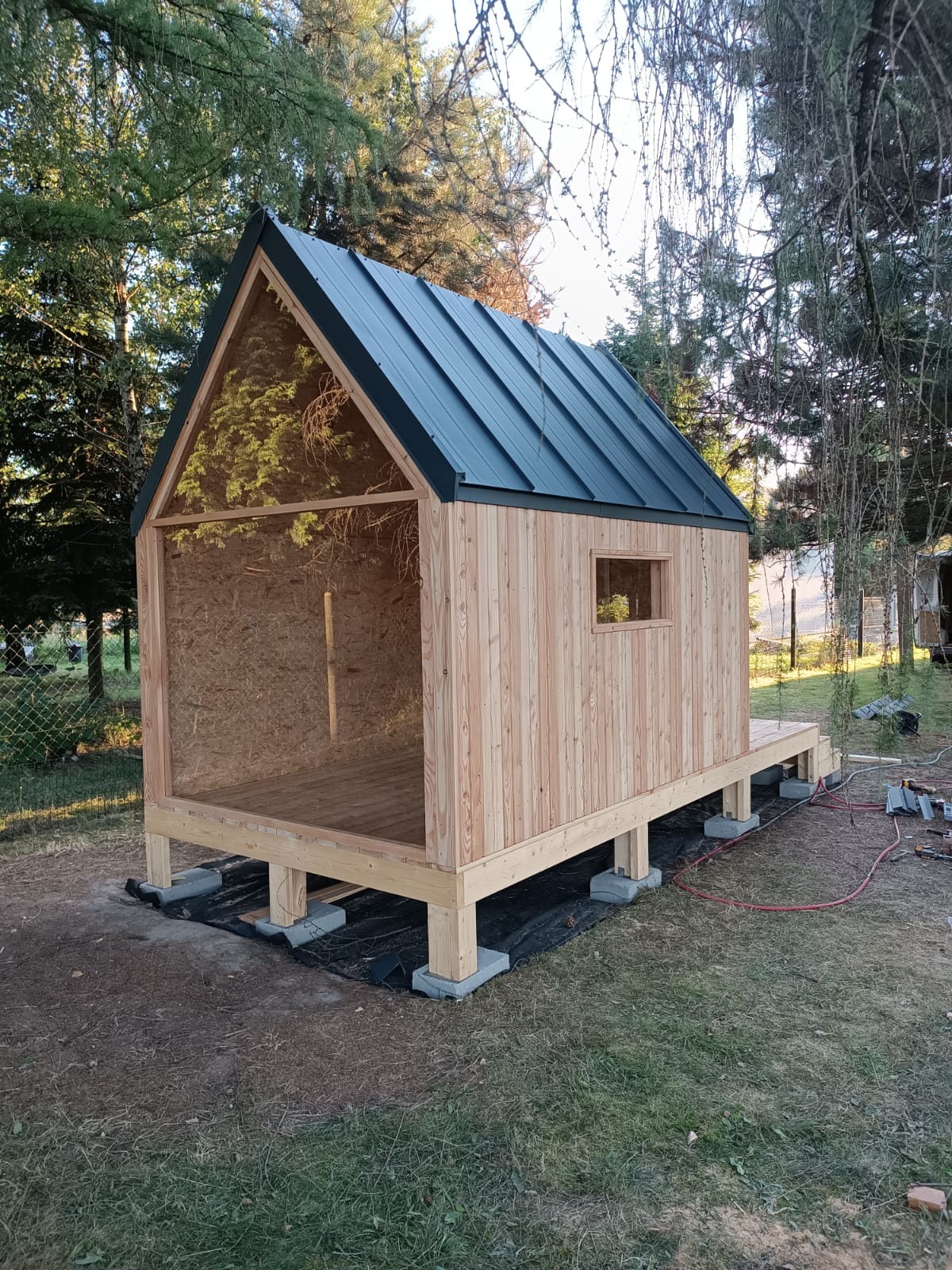 Surowa konstrukcja drewniana małego domku z dachem z blachy na rąbek, posadowiona na betonowych bloczkach, widoczne narzędzia i przewody elektryczne sugerują etap budowy.
