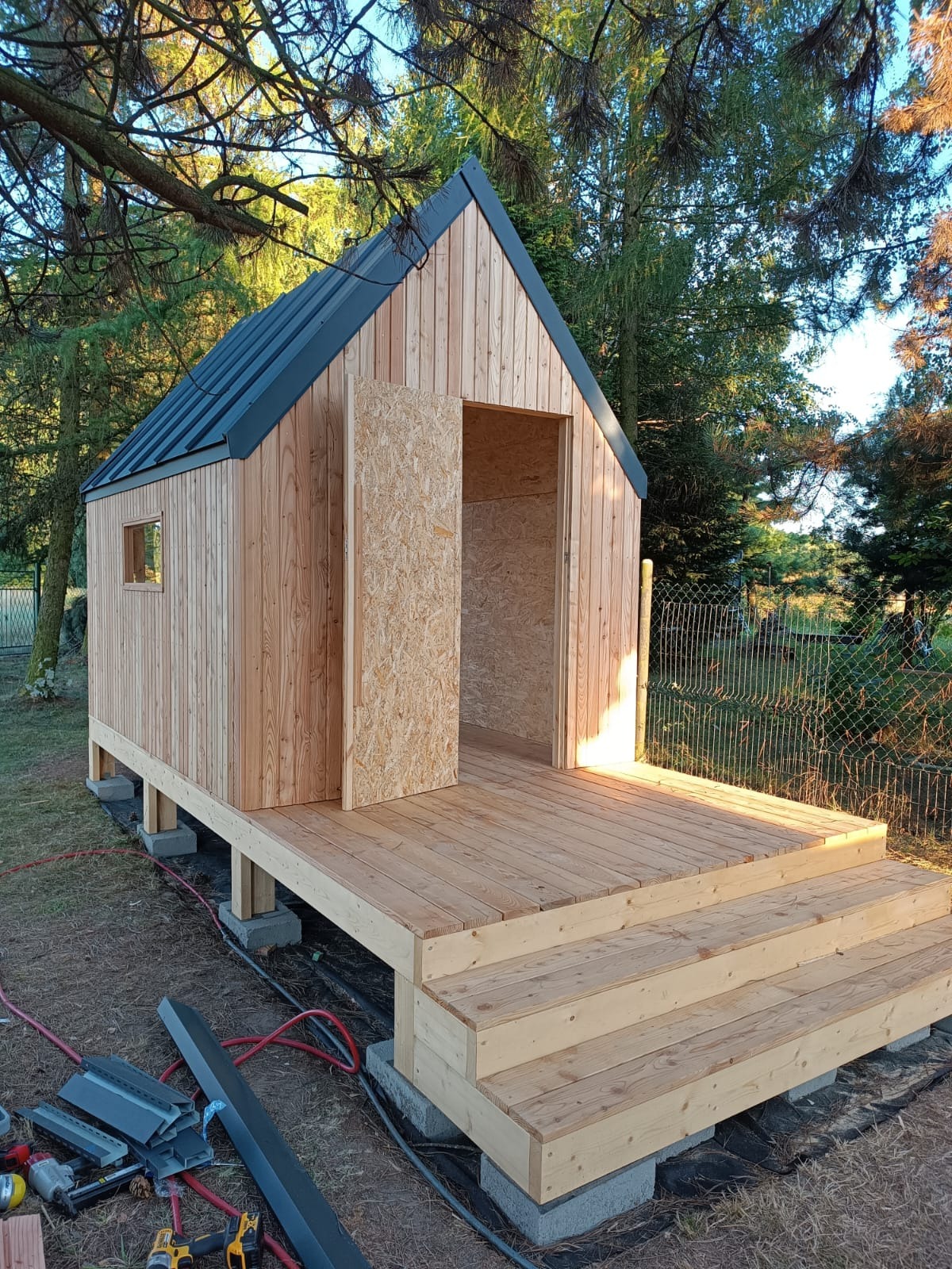 Surowy, drewniany domek z czarnym dachem, posadowiony na betonowych bloczkach, z widocznymi schodami wejściowymi i częściowo zamontowanymi drzwiami z płyty OSB, otoczony zielenią i narzędziami...