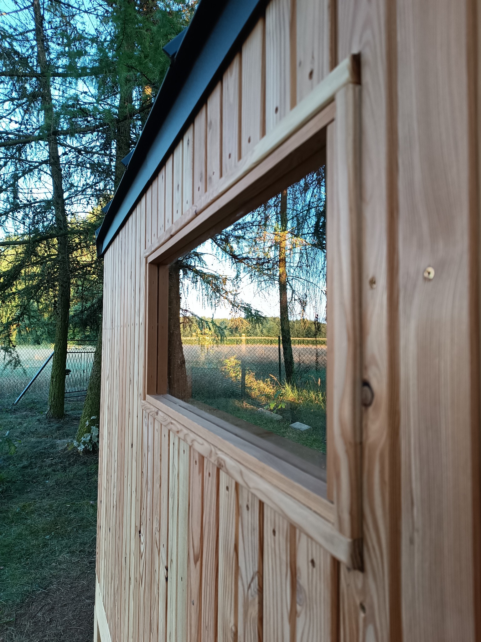 Fragment ściany zewnętrznej domu z drewnianych paneli z widocznym oknem, w którym odbija się krajobraz z drzewami i polem.