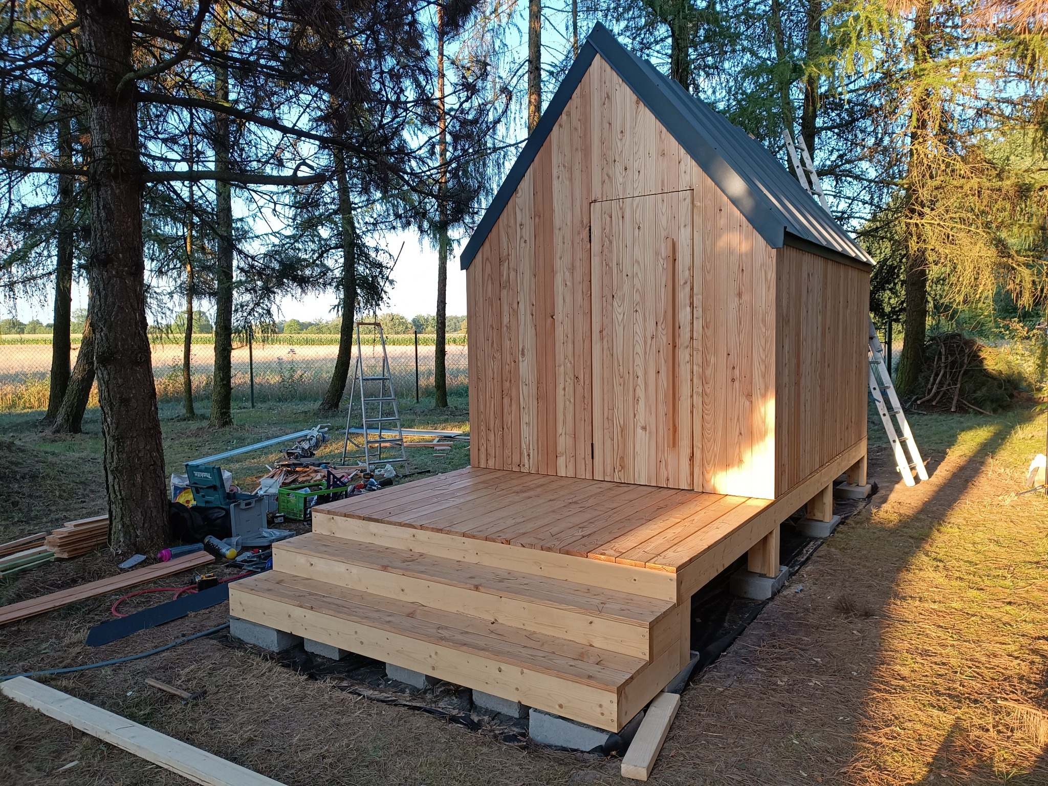 Niewielki, drewniany budynek z dwuspadowym dachem, posadowiony na betonowych bloczkach, z dobudowanym tarasem i schodami wejściowymi, otoczony drzewami i narzędziami budowlanymi.