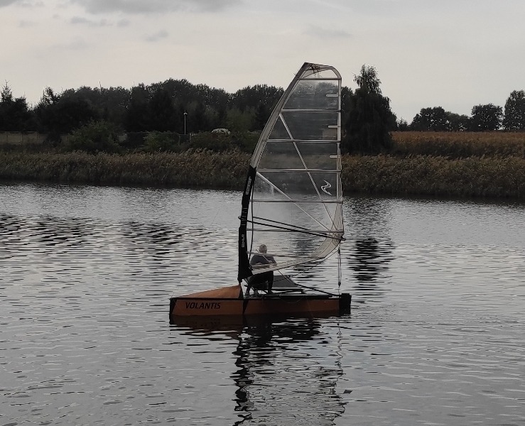 Mała żaglówka Volantis na spokojnej wodzie, z widocznym żaglem i siedzącą osobą, otoczona trzcinami i drzewami w pochmurny dzień.