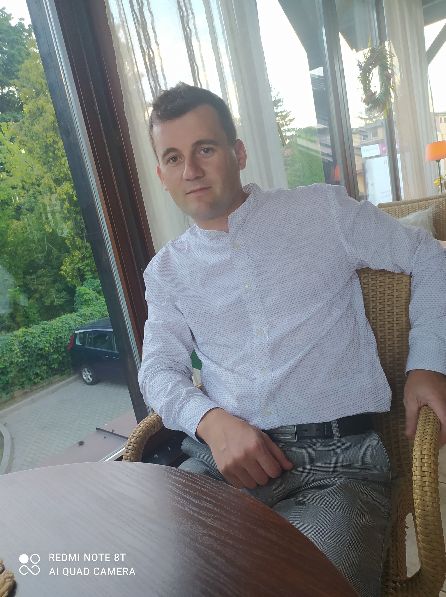 Mężczyzna w koszuli siedzi w wiklinowym fotelu przy oknie z widokiem na zieleń i samochód. W tle budynki i dekoracyjny wieniec.