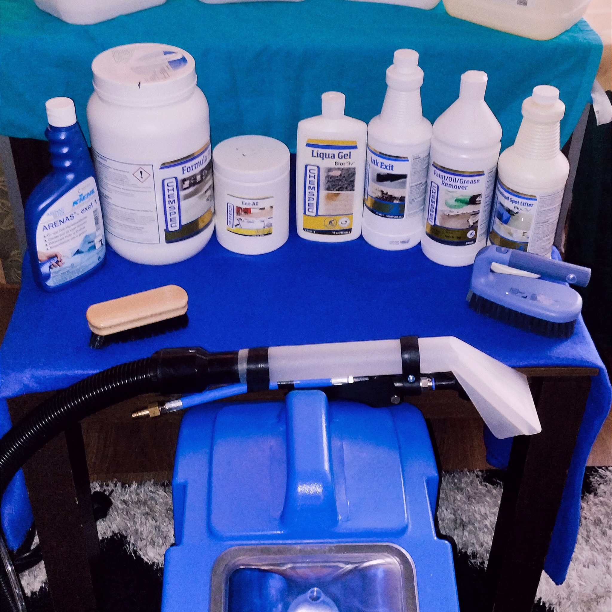 Zestaw środków czyszczących i akcesoriów do czyszczenia tapicerki, w tym szczotki i odkurzacz ekstrakcyjny, prezentowany na niebieskim tle.