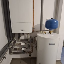 INSTAL-HEAT RADOSŁAW MORAWSKI - Biały kondensacyjny kocioł gazowy marki Junkers oraz biały zasobnik ciepłej wody użytkowej marki Bosch z niebieskim naczyniem przeponowym, połączone rurami miedzianymi i szarymi rurami z izolacją.