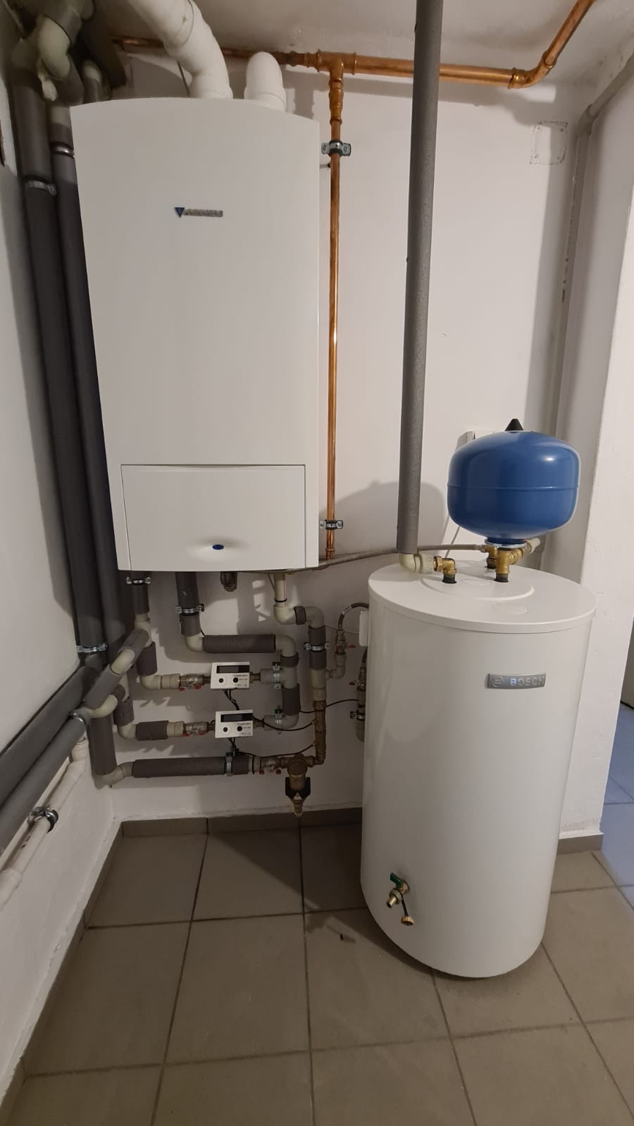 Biały kondensacyjny kocioł gazowy marki Junkers oraz biały zasobnik ciepłej wody użytkowej marki Bosch z niebieskim naczyniem przeponowym, połączone rurami miedzianymi i szarymi rurami z izolacją.