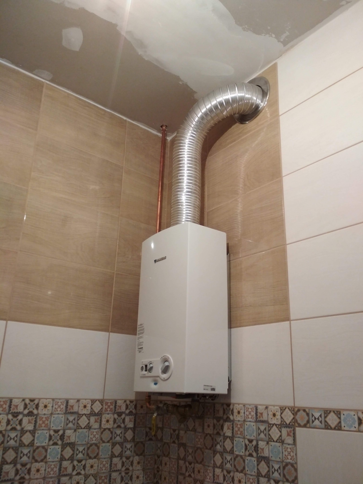 Zamontowany, biały piec gazowy marki Junkers z podłączonym kominem aluminiowym w pomieszczeniu wyłożonym płytkami ceramicznymi o różnych wzorach i kolorach.