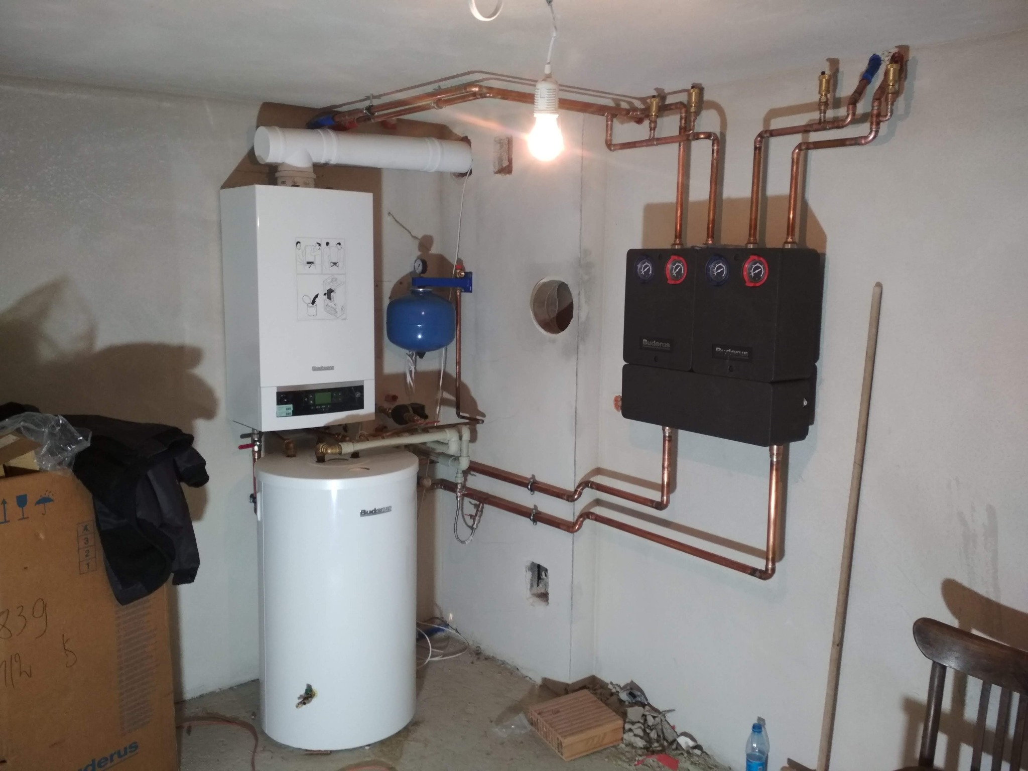 Zainstalowany, nowoczesny piec gazowy dwufunkcyjny Buderus z zasobnikiem i osprzętem, widoczne miedziane rury instalacji grzewczej, całość w surowym pomieszczeniu z betonową podłogą i gołą żarówką.