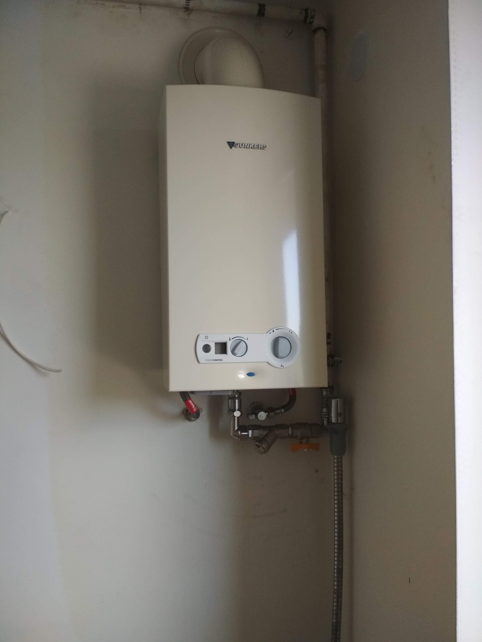 Biały piec gazowy Junkers zamontowany na ścianie, widoczne rury i zawory instalacji gazowej, otoczenie to jasne ściany.