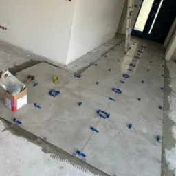 Układanie szarych płytek podłogowych z użyciem niebieskich klipsów poziomujących, widoczna drabina oparta o ścianę i drzwi zabezpieczone folią, karton z zaprawą klejową i gumowy młotek.